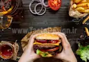 یک ساندویچ همبرگر در دستان یک فرد در کنار نوشابه و سیب زمینی